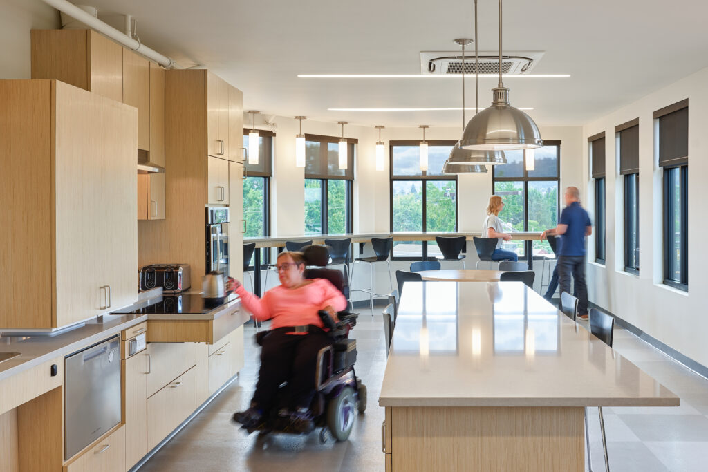 A person in a peach-colored shirt who uses a wheelchair, takes a kettle off a induction cooktop. The spacious kitchen features many accessible designs. People stand and chat in the background.