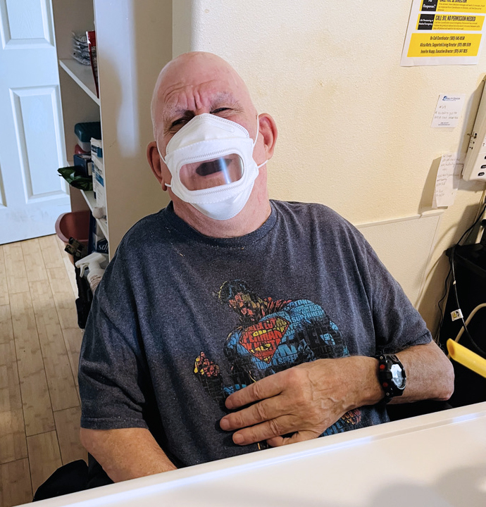 Older man in PPE mask is seated at a table with an impish look in his eyes