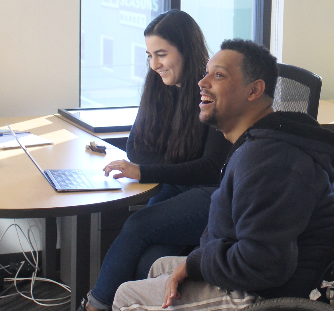 Two smiling people sitting at a desk working on a laptop