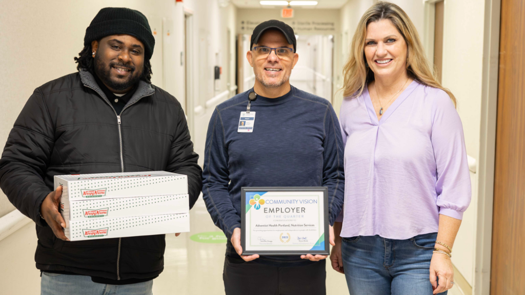 Three smiling people stand in a hospital hallway. One of them holds a framed certificate, another holds three boxes of donuts.