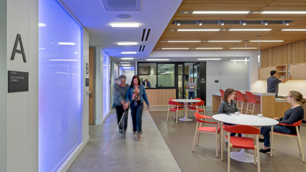 Two people walking through a wide corridor with tables on one side and offices on the other. One of the people uses a cane to navigate. Image credit: Lighthouse for the Blind in San Francisco, Mark Cavagnero Associates; photo by Jasper Sanidad. https://www.cavagnero.com/project/lighthouse-for-the-blind-and-visually-impaired/