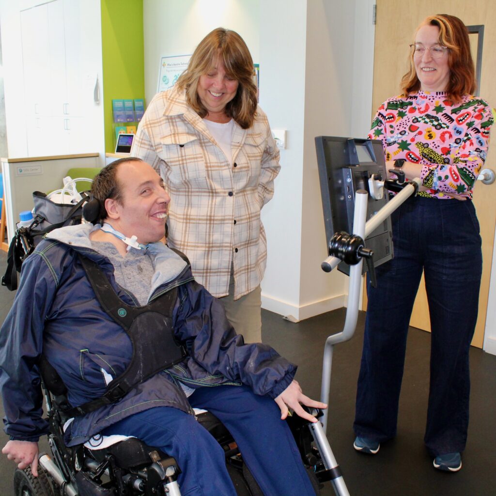 A handsome man who uses AAC and a wheelchair smiles at the camera while two woman look at him and smile