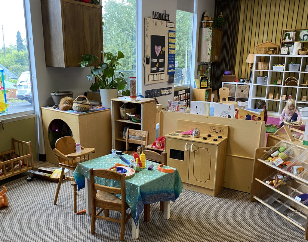 A warm and inviting preschool room with a small play kitchen and table in the foreground. Two children play on a rug in the background