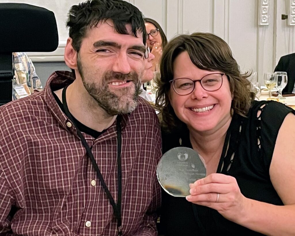 Smiling man and smiling woman hold up shiny, circle-shaped award