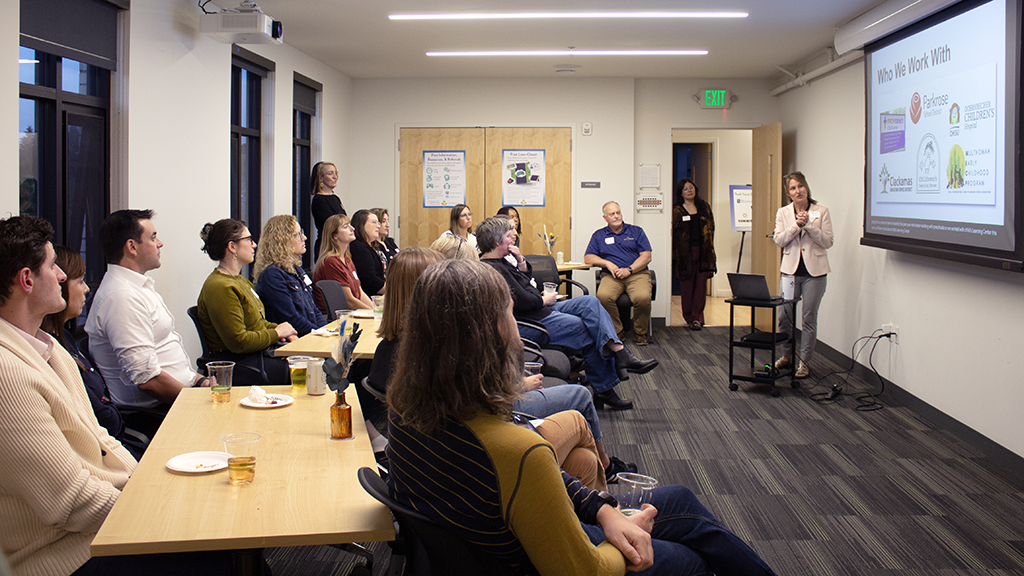 A woman gives a presentation to about 20 people seated around tables in a conference room