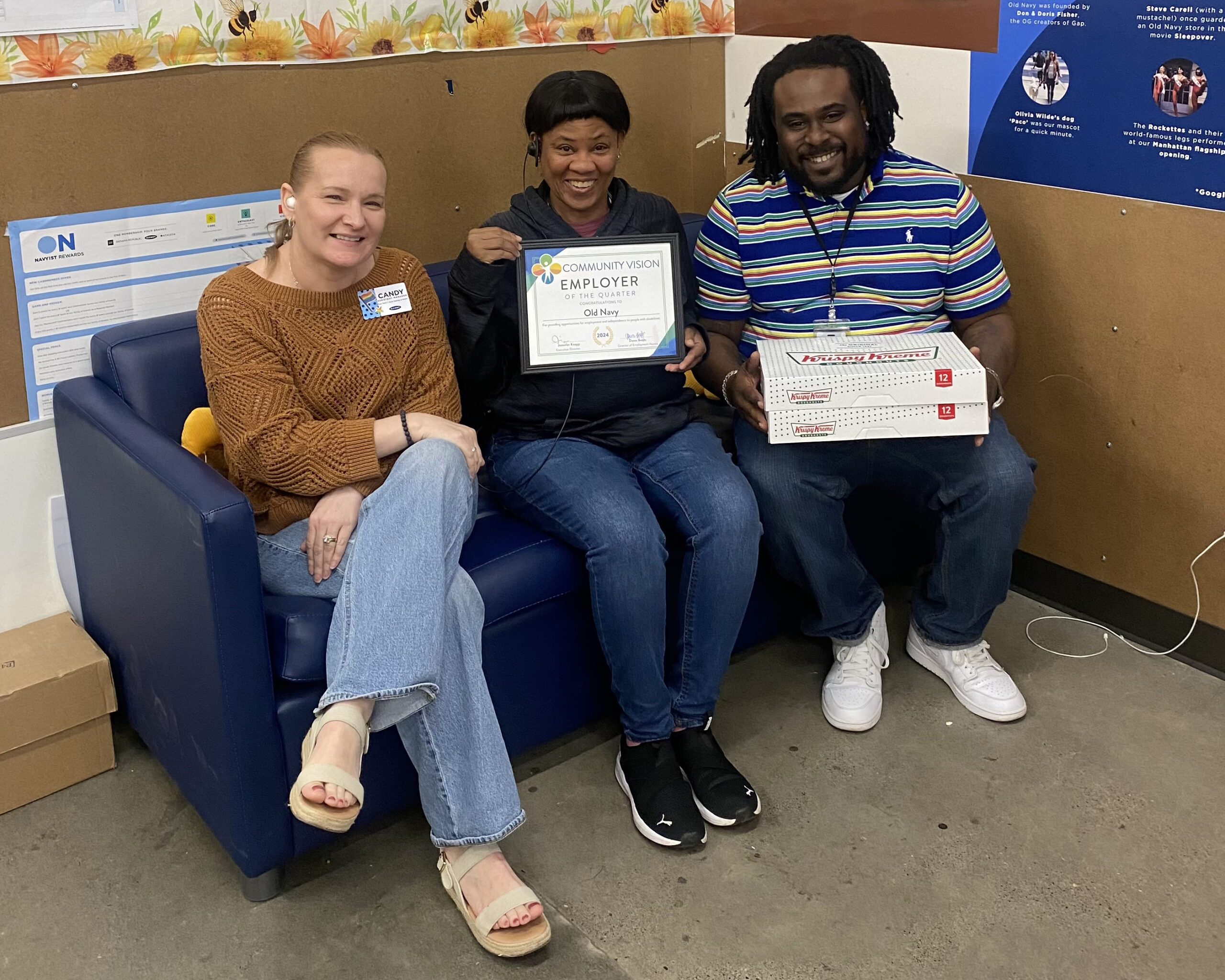 Three people smile and sit on a blue couch in a break room. One holds a certificate. Another holds 2 boxes of doughnuts.