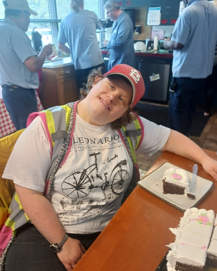 Smiling woman in safety vest and cap sits at a table with a piece of birthday cake