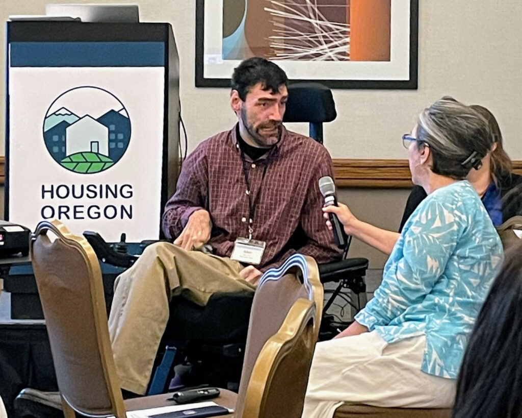 Man who uses wheelchair speaks into microphone with "Housing Oregon" poster behind him