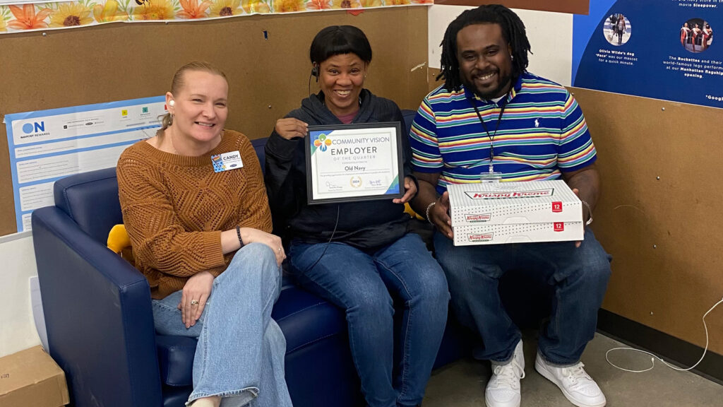 Three people smile and sit on a blue couch in a break room. One holds a certificate. Another holds 2 boxes of doughnuts.