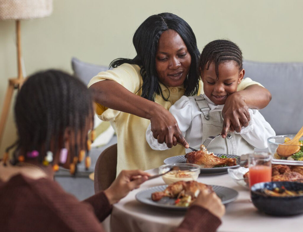 A child sits on their mother's lap at a holiday meal. The mother helps cut the child's food. A sibling looks on.