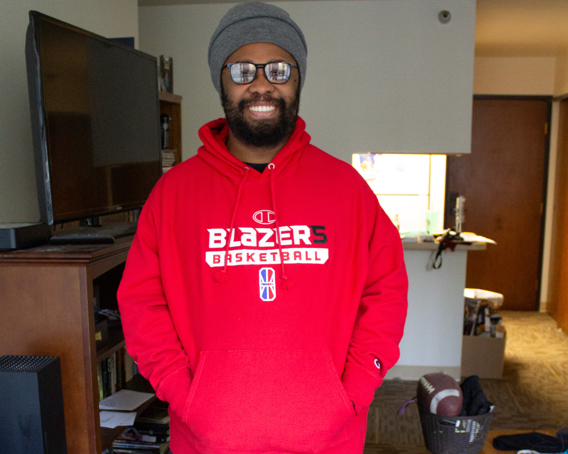 A young man with a beard and glasses stands in his apartment with a TV and kitchen in the background