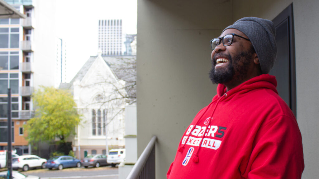 A man with a beard and glasses stands on a balcony and looks upward while smiling