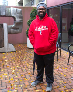 A young man with a beard and glasses, wearing a red hoodie and grey beanie, stands outside on a brick patio with a metal sculpture behind him