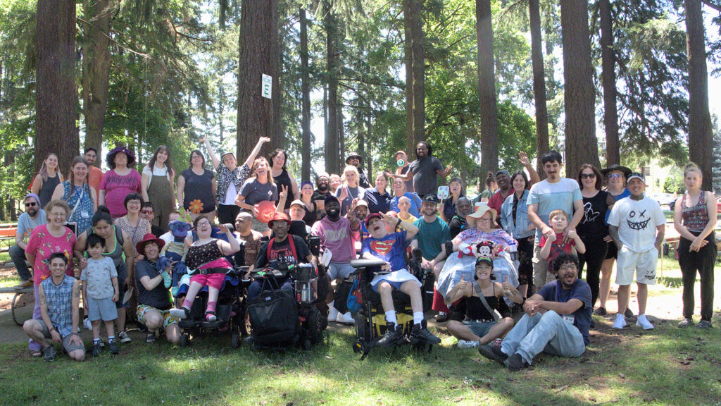 Group photo of about 40 people standing in a park with sun-dappled Douglas Fir trees in the background