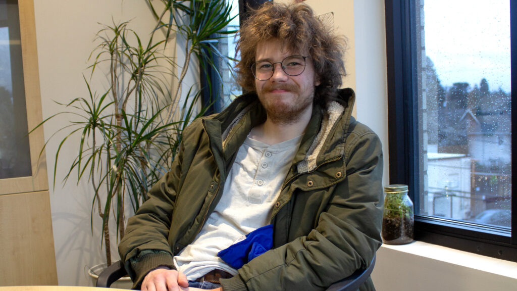 Young man who wears glasses and has a scruffy brown beard and brown hair sits in a chair in front of windows on a raining day and smiles