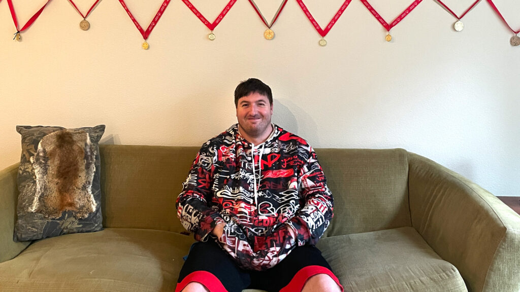 Man in red, black, and white print hoody sits on a couch under row of medals hanging above his head