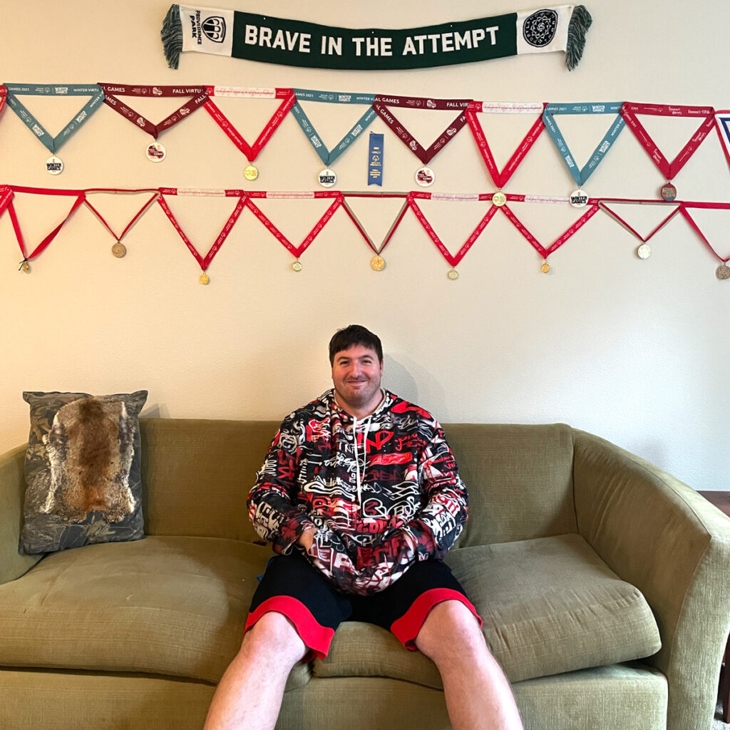 Man in red, black, and white print hoody sits on a couch under two rows of medals hanging above his head