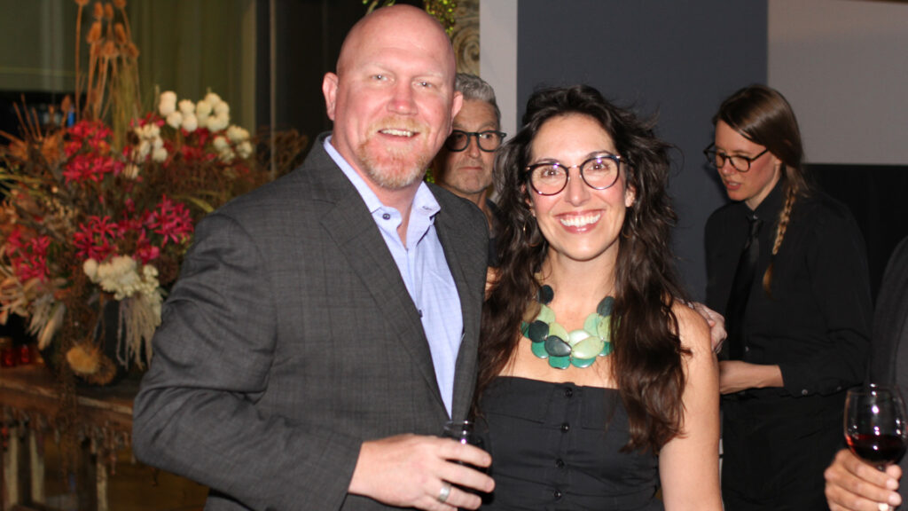 A man in a sport coat holding a glass of wine stands next to a woman in glasses and a large green necklace