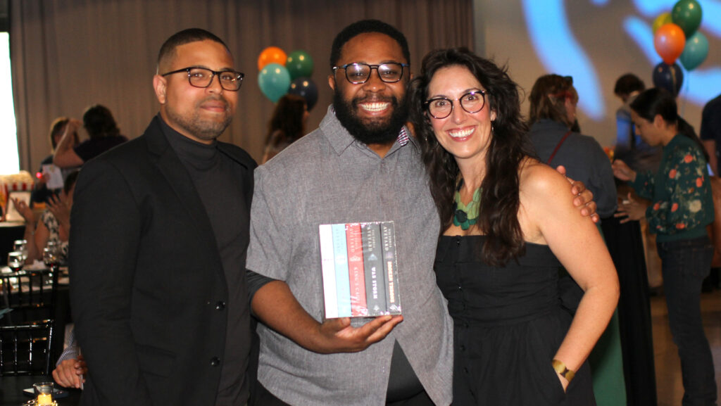 Three people pose for a photo. The man in the middle smiles widely and holds a box set of books