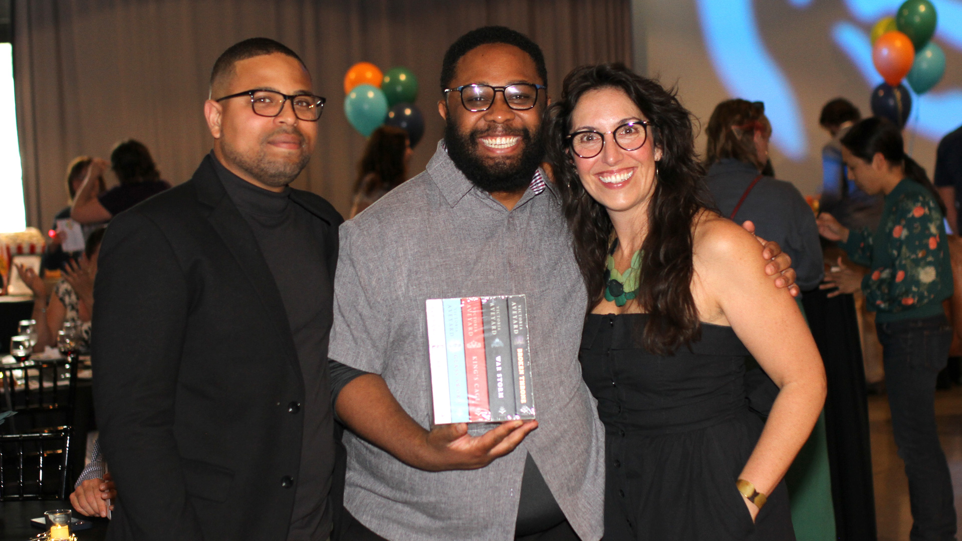 Three people pose for a photo. The man in the middle smiles widely and holds a box set of books