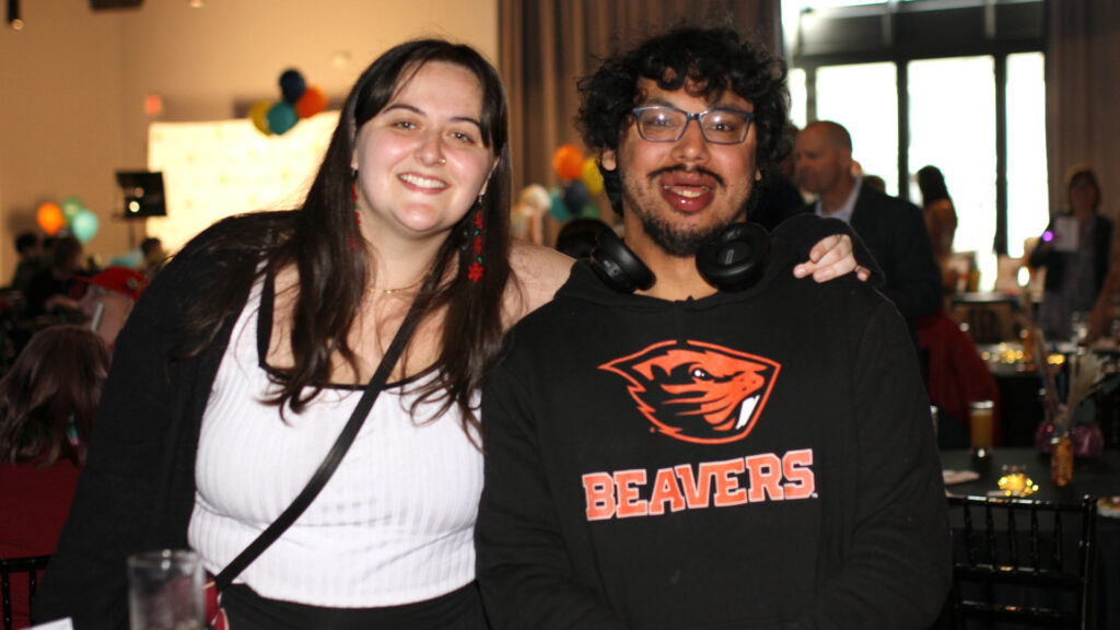 A young man in headphones and a Beavers sweatshirt smiles next to a woman with long hair and dangly earrings