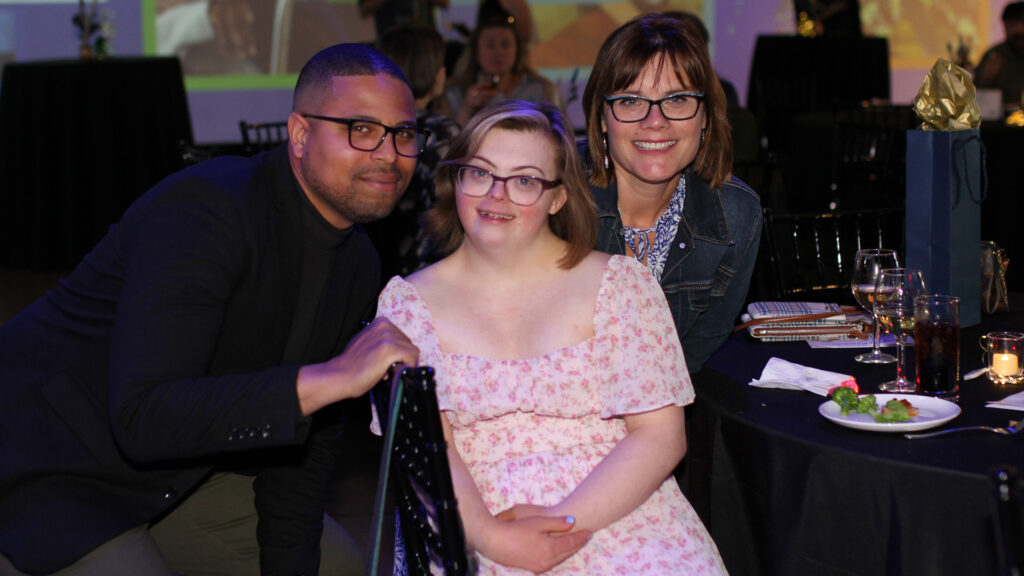 A smiling woman in a pink print dress and glasses poses for a photo at a gala event with two friends on either side of her