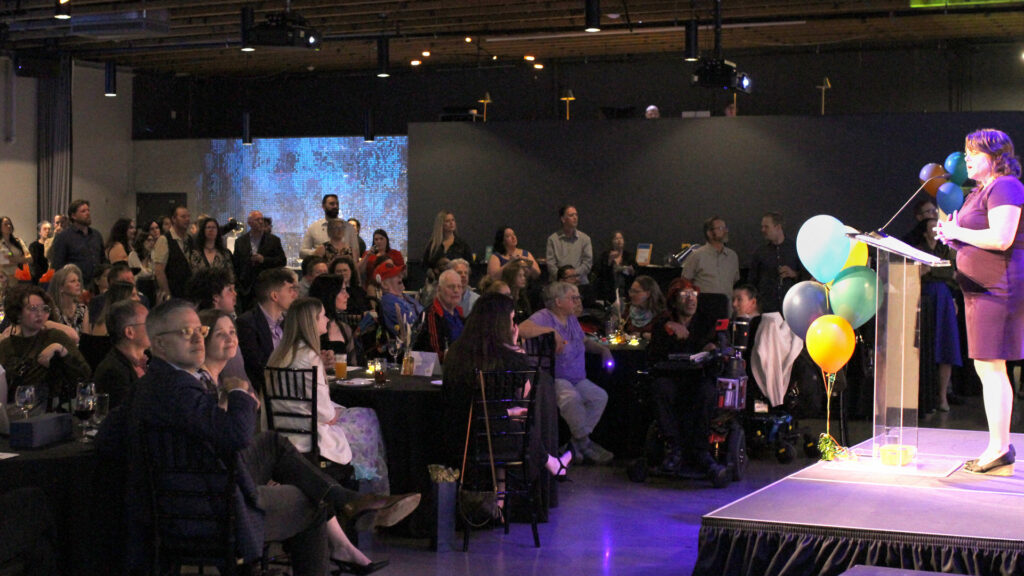 An audience sits at tables and stands in a darkened room and listen to a woman at a podium with balloons