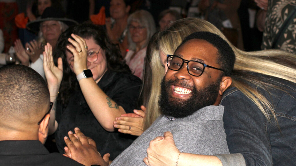 Man with glasses and disarming smile is hugged by woman as others around him applaud