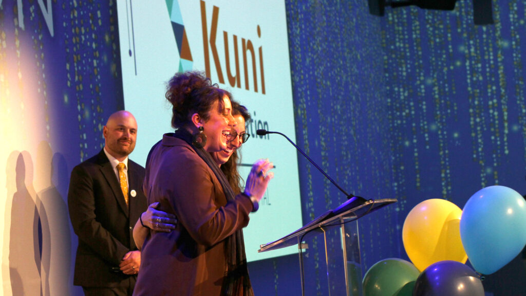 Three people on a stage smiling with balloons and the Kuni Foundation logo in the background
