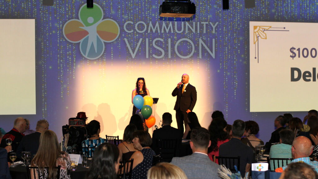 Two people stand at a podium with balloons and the Community Vision logo in the background