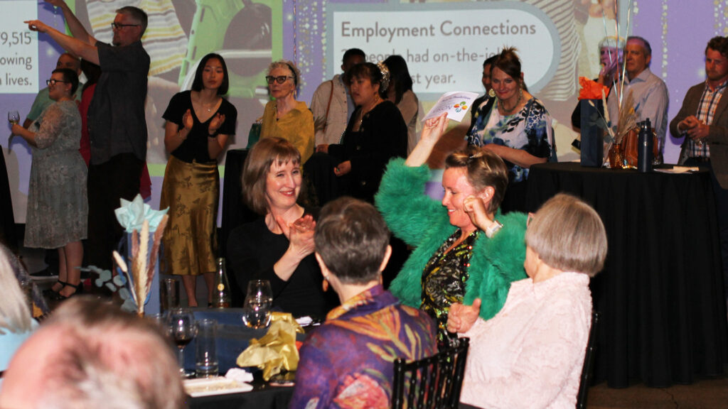 Woman in shaggy green coat and sparkly dress celebrates and holds up a bid number