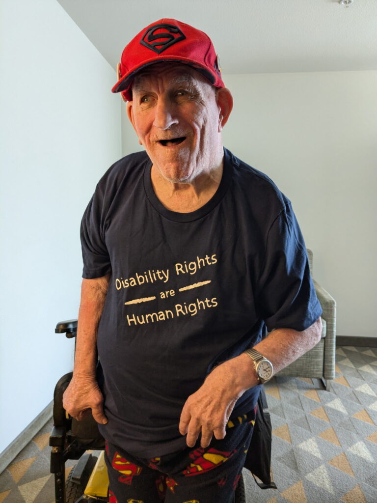 An older man in a red Superman cap and a t-shirt that says "Disability Rights Are Human Rights" smiles for a photo