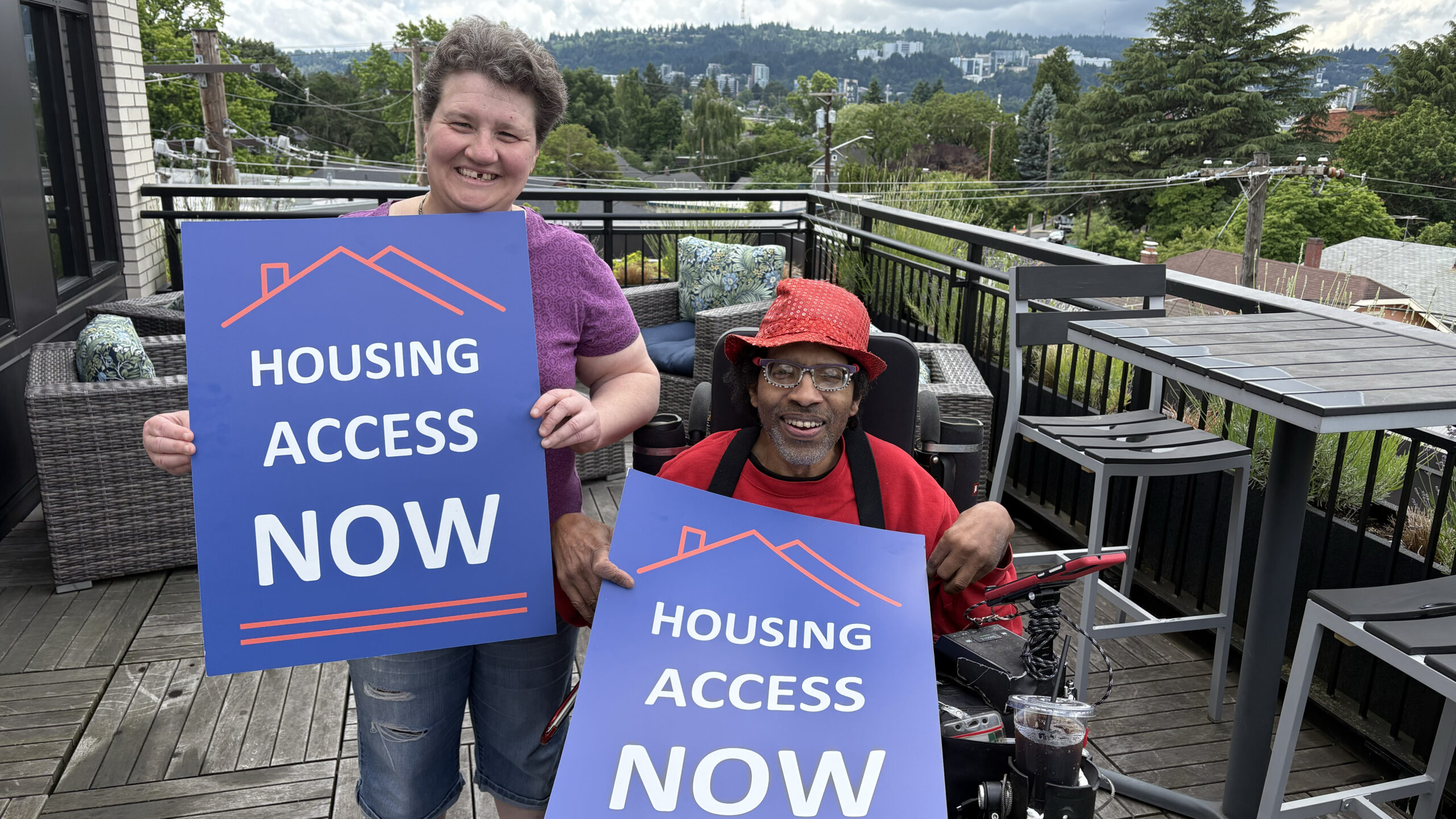A person with short wavy hair and a cute smile stands next to a person wearing a bright red hat, sitting in a a wheelchair. They hold up rally signs that say "Housing Access Now."