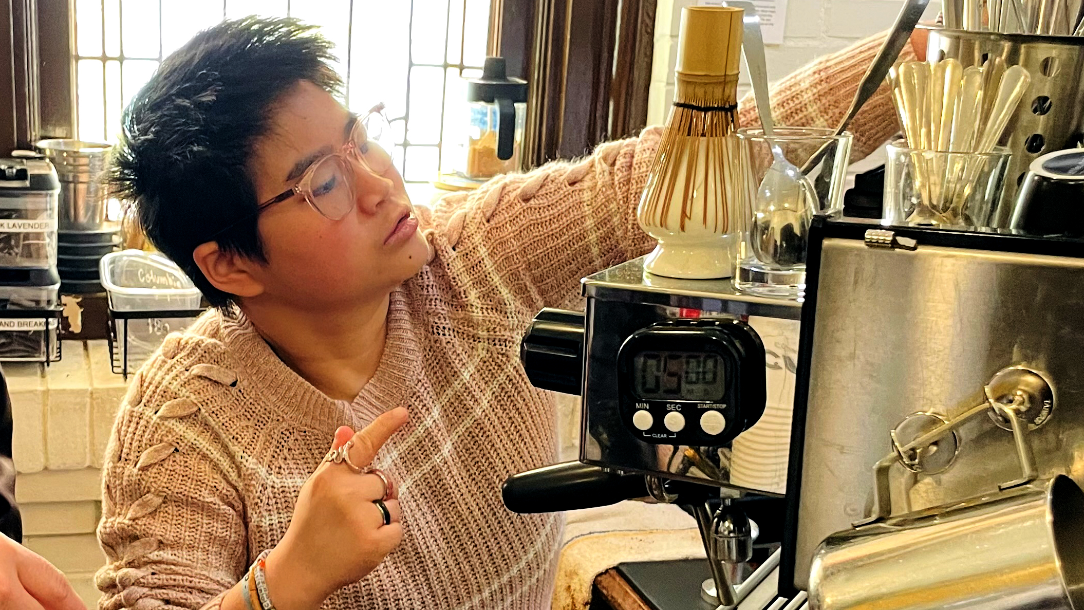 A women with cute short hair and glasses stands behind a counter and sets something on top of an espresso machine. A leaded glass window lets in day light behind her.