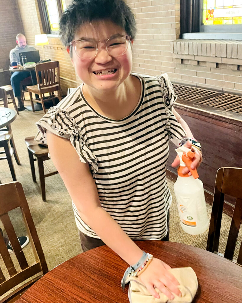 A smiling women with short, dark hair and glasses and wearing a cute striped top, cleans a table in a cafe.