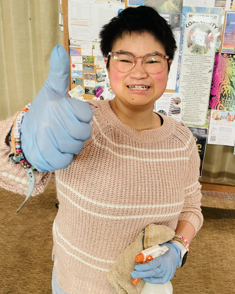 A smiling women with short, dark hair and glasses gives a thumbs up with one hand and holds a cleaning spray bottle in the other.