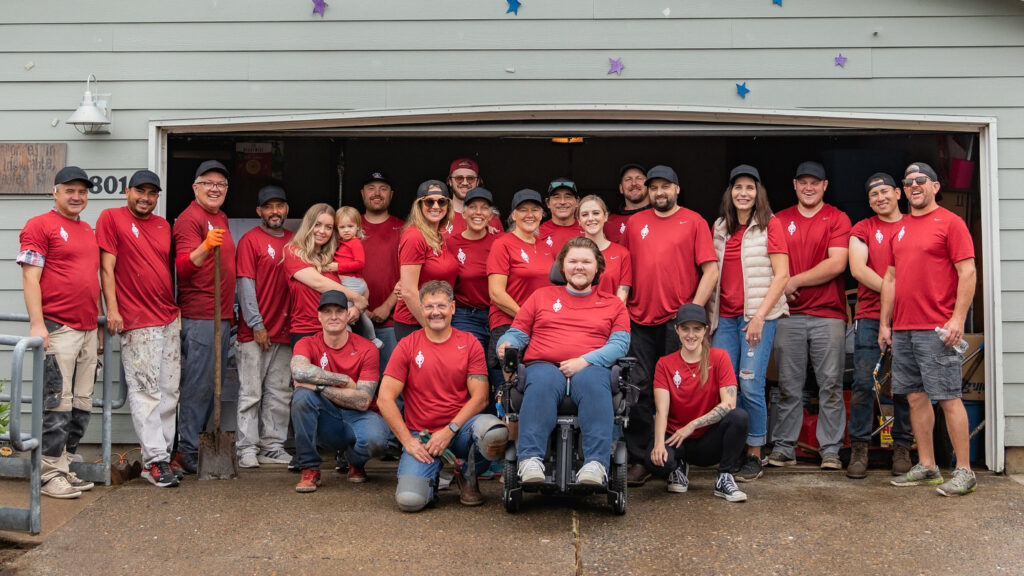 Two dozen people in red t-shirts pose for a group photo in front of a garage