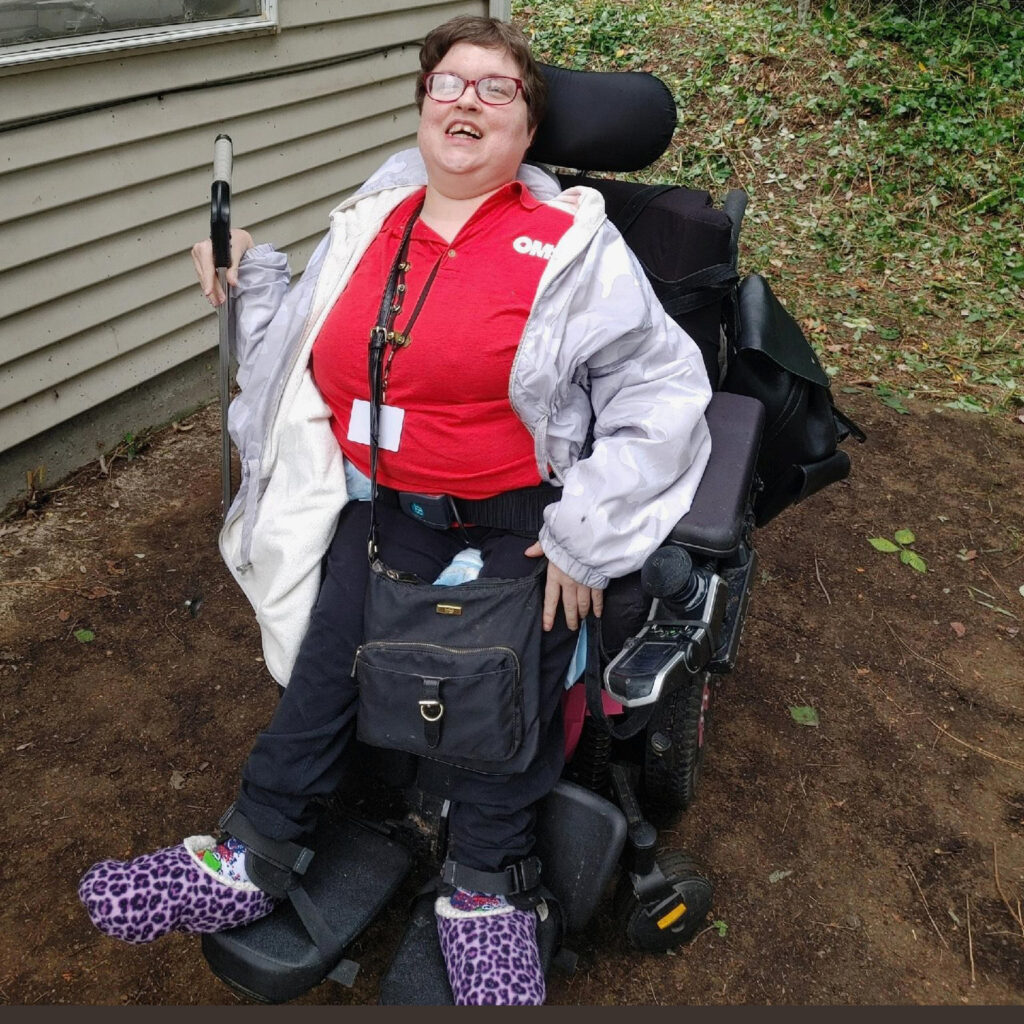 A smiling woman in a red t-shirt, sitting in a wheel chair, is in a cleared backyard
