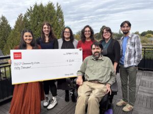 A group of smiling people, including one person in a wheel chair, hold up a big check for $50,000 with a Wells Fargo logo on it