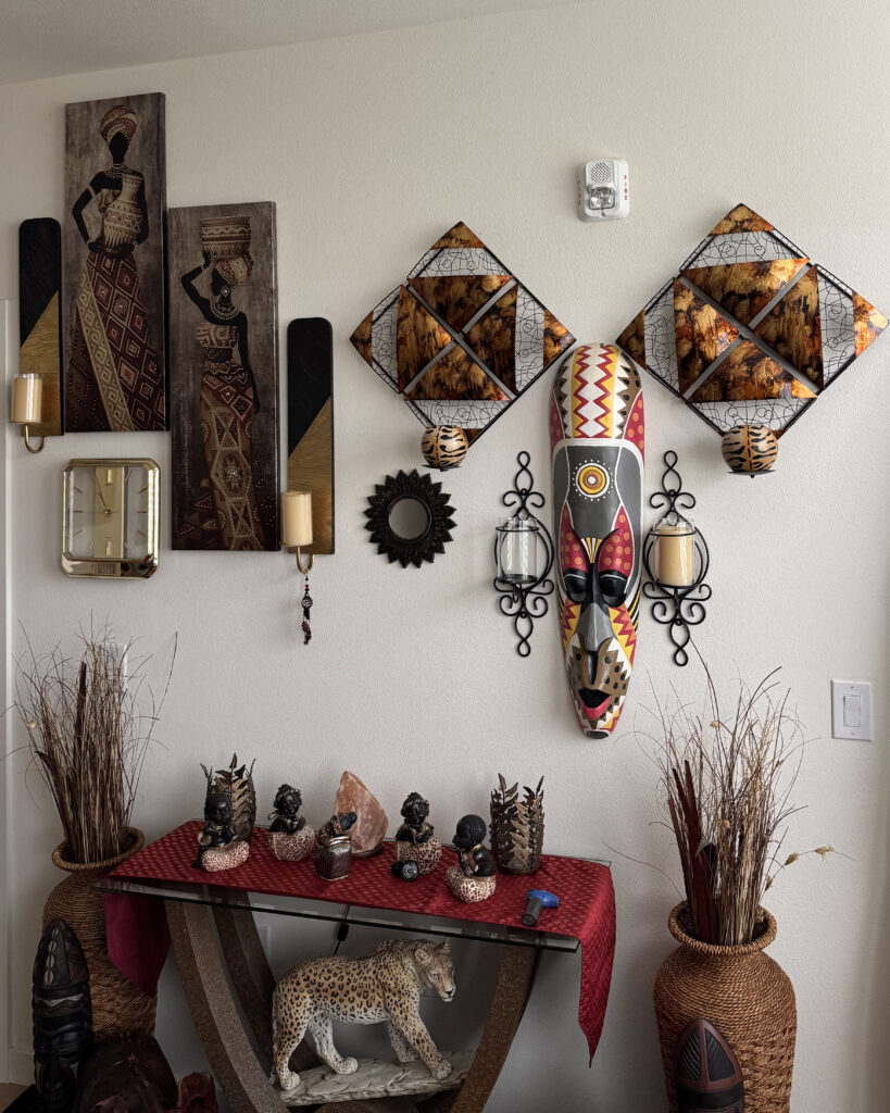 An apartment interior wall carefully decorated with artwork, a mask, a clock, sconces, dried plants, statutes, and crystals. The color theme is earth tones with red accents.