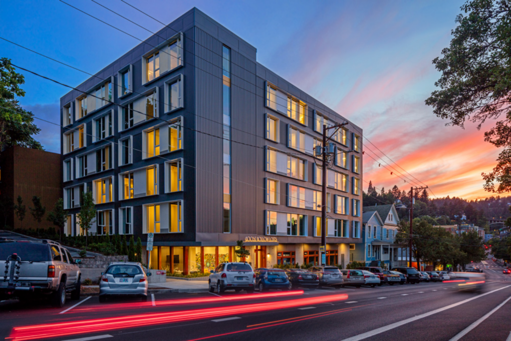 A modern apartment building with many windows at dusk, cars parked in front.