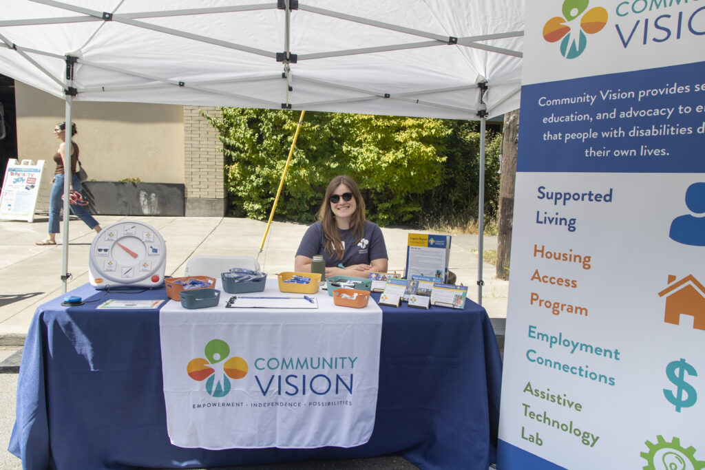 Woman with long hair wearing a blue t-shirt and black sunglasses smiles while sitting at a table in a booth with Community Vision signs.