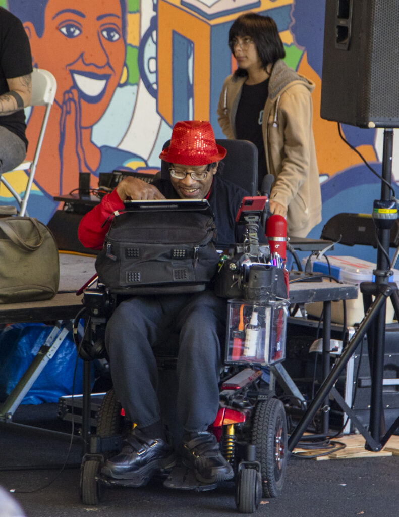 _Z3A9652 A man in a power wheelchair wearing a red hat and glasses smiles and taps a tablet next to a stage.