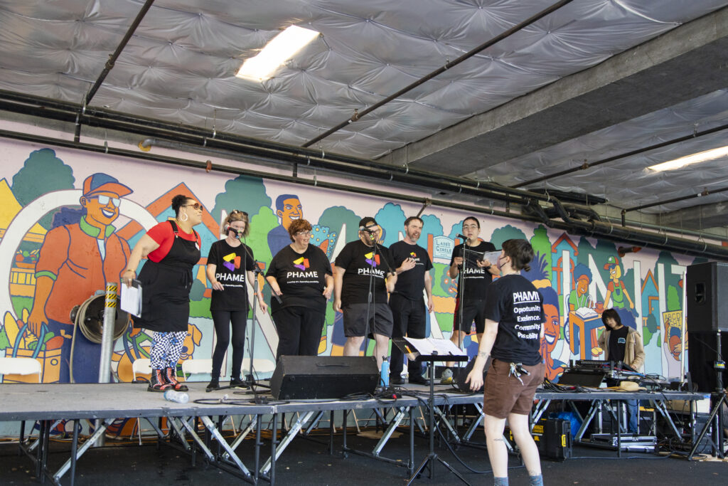 _Z3A9923 A group of 6 people wearing black t-shirts singing on a stage. One person in a black t-shirt and brown shorts stands in front of them directing.